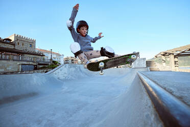Teen boy in protective helmet riding skateboard in skate park on sunny day on seashore - ADSF29640