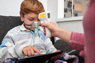 Unerkennbar liebende Mutter mit Infrarot-Thermometer, die die Temperatur eines Jungen misst, der während einer Inhalation in einer Sauerstoffmaske atmet und sich einen Zeichentrickfilm auf einem Tablet zu Hause ansieht - ADSF29648
