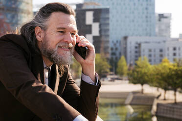 Side view of smiling bearded mature male with gray hair wearing classy suit discussing business issues during phone conversation while standing against modern city buildings - ADSF29657