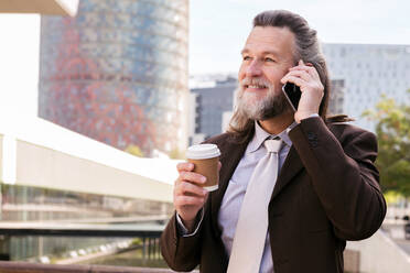 Smiling mature bearded man in stylish elegant suit with cup of takeaway coffee in hand speaking on mobile phone while standing on urban street - ADSF29659