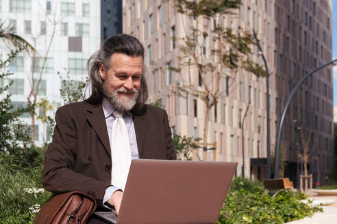 Glücklicher, erfolgreicher, grauhaariger, bärtiger Mann im eleganten Anzug, der einen Laptop benutzt, während er auf einer städtischen Straße sitzt - ADSF29665