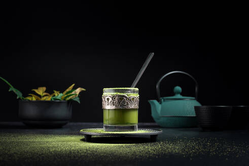 Still life composition with traditional oriental matcha tea served in glass cup with metal ornamental decor on table with ceramic bowls and fresh green leaves against black background - ADSF29675