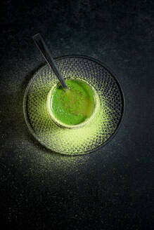 From above of healthy herbal green matcha tea served in glass cup with metal decoration on saucer sprinkled with powder on black table - ADSF29676