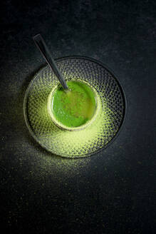 Von oben gesunder grüner Matcha-Kräutertee, serviert in einer Glastasse mit Metalldekoration auf einer Untertasse, bestreut mit Pulver auf einem schwarzen Tisch - ADSF29676
