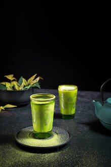 Still life composition with traditional oriental matcha tea served in glasses cups with metal ornamental decor on table with ceramic bowls and fresh green leaves against black background - ADSF29679