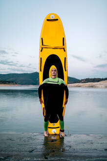 Ruhiger männlicher Surfer im Neoprenanzug stehend, Augen geschlossen, mit gelbem SUP-Board am Strand in der Nähe des Meeres - ADSF29683