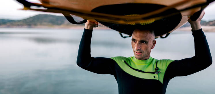 Älterer männlicher Surfer steht mit Paddleboard über dem Kopf in der Nähe des Meeres am Abend und schaut weg - ADSF29684