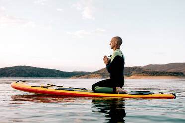 Seitenansicht eines ruhigen männlichen Surfers, der in Thunderbolt-Pose mit Namaste-Händen auf einem Paddelbrett sitzt und meditiert, während er abends im Meer Yoga übt - ADSF29687