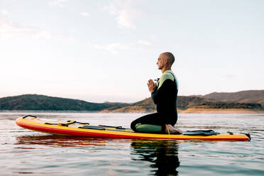Side view of tranquil male surfer sitting in Thunderbolt pose with Namaste hands on paddleboard and mediating while practicing yoga in sea in evening - ADSF29687
