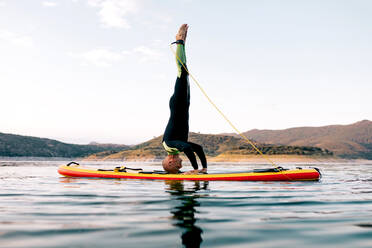 Seitenansicht eines männlichen Surfers, der Yoga im Kopfstand auf einem SUP-Board im Meer bei Sonnenuntergang praktiziert - ADSF29688