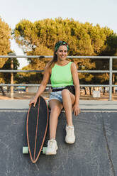 Lächelnde Frau mit Skateboard im Park sitzend - JRVF01759