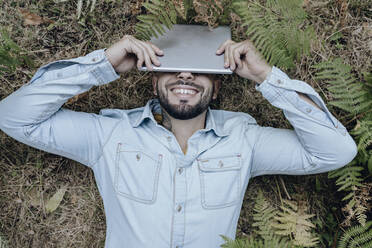 Carefree man lying on plants while covering face with digital tablet in forest - JCCMF03801