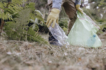 Männlicher Freiwilliger mit Handschuh, der beim Säubern des Waldes eine Plastikflasche aufhebt - JCCMF03810