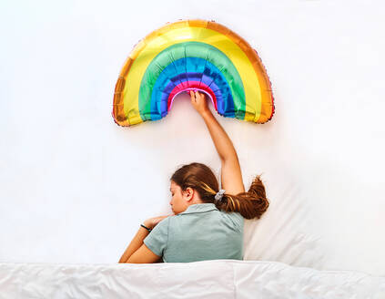 Draufsicht auf eine junge Frau, die einen mehrfarbigen aufblasbaren Regenbogenballon hält, während sie unter einer Decke in einem Bett mit weißen Laken schläft - ADSF29718