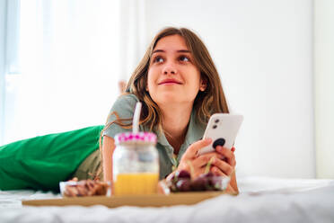 Junge Studentin beim Surfen in sozialen Netzwerken auf dem Handy in der Nähe eines Tisches mit frischem Obst und Saft, während sie den Morgen zu Hause verbringt - ADSF29721