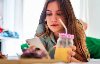Junge Studentin beim Surfen in sozialen Netzwerken auf dem Handy in der Nähe eines Tisches mit frischem Obst und Saft, während sie den Morgen zu Hause verbringt - ADSF29722