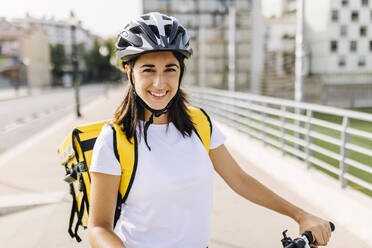 Lächelnde Servicefrau mit Fahrradhelm und Lieferrucksack an einem sonnigen Tag - XLGF02237