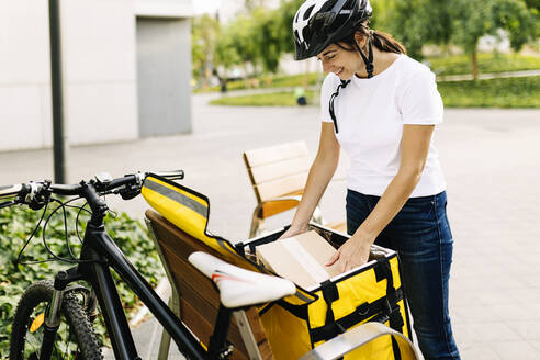 Zustellerin, die auf einer Bank mit dem Fahrrad einen Kurier aus dem Rucksack nimmt - XLGF02262