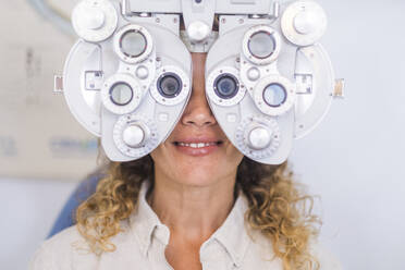 Smiling female patient examining eyesight through phoropter in medical clinic - SIPF02346