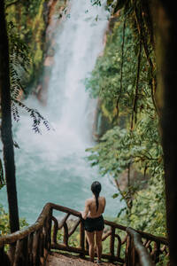Von oben Rückenansicht einer nicht erkennbaren weiblichen Touristin, die auf einem Aussichtspunkt steht und die malerische Landschaft mit dem plätschernden Wasserfall und dem türkisfarbenen Wasser des Celeste-Flusses inmitten des üppigen grünen Laubes in Costa Rica betrachtet - ADSF29747