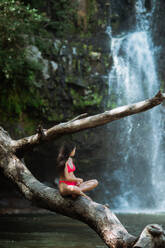 Side view of unrecognizable slim female traveler in pink bikini sitting on large old tree branch against picturesque waterfall cascade falling from rocky slope in summer day in Costa Rica - ADSF29751