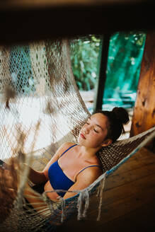 Side view of serene young female traveler in bikini chilling in hammock hanging on terrace of tropical house during summer holidays in Uvita town in Costa Rica - ADSF29754