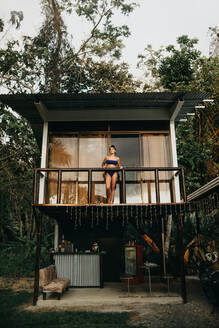 From below full body of young long haired female traveler in stylish swimwear standing on balcony of beach house located near green trees in summer day in Uvita town in Costa Rica - ADSF29760
