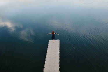 High angle back view of female standing with outstretched arms on quay near lake and enjoying freedom - ADSF29763