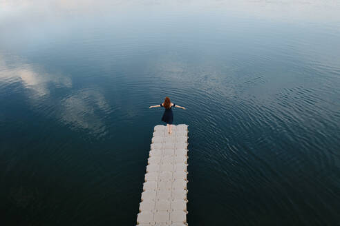 Hochformatige Rückenansicht einer Frau, die mit ausgestreckten Armen auf einem Kai in der Nähe eines Sees steht und die Freiheit genießt - ADSF29763