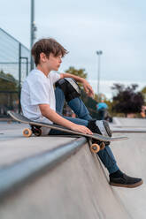 Side view of teen boy sitting with skateboard on ramp in skate park and looking away - ADSF29768