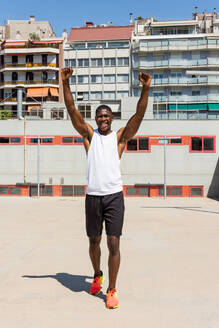 Positiver afroamerikanischer männlicher Sportler in Sportkleidung, der mit erhobenen Armen auf einem Sportplatz steht und seine Leistung feiert, während er in die Kamera schaut - ADSF29769