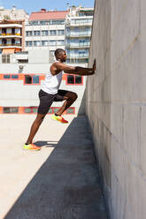 Seitenansicht eines athletischen afroamerikanischen Mannes, der sich an die Wand lehnt und auf der Stelle läuft, während er an einem sonnigen Tag auf einem Sportplatz aktiv trainiert - ADSF29770