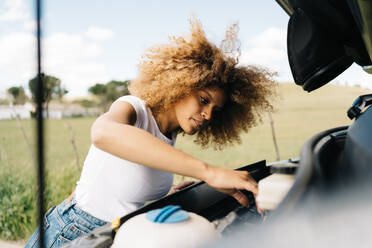 Seitenansicht einer reisenden Frau, die unter die geöffnete Motorhaube eines Wohnmobils schaut, während sie während einer Reise durch die sommerliche Natur Probleme hat - ADSF29776