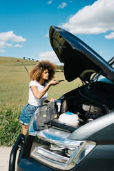 Junge ethnische Frau mit Afro-Haar, die mit ihrem Smartphone einen Reparaturdienst anruft, während sie in der Nähe eines Wohnmobils mit offenem Verdeck auf einer Landstraße im Sommer steht - ADSF29778
