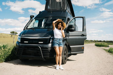 Worried young African American female with curly hair talking on mobile phone while asking for help from repair service after accident with camper van in countryside - ADSF29779