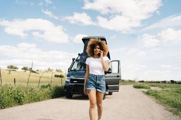 Worried young African American female with curly hair talking on mobile phone while asking for help from repair service after accident with camper van in countryside - ADSF29780