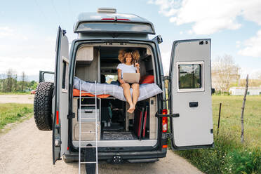 Junge afroamerikanische Reisende mit lockigem Haar, die Bier trinkt und einen Film auf dem Laptop anschaut, während sie sich im Wohnmobil während der Sommerferien ausruht - ADSF29784