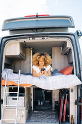 Glückliche Afroamerikanerin mit lockigem Haar, die lächelt und auf ihrem Handy surft, während sie sich auf dem Bett eines Wohnwagens ausruht - ADSF29787