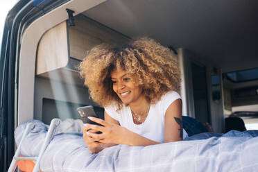 Glückliche Afroamerikanerin mit lockigem Haar, die lächelt und auf ihrem Handy surft, während sie sich auf dem Bett eines Wohnwagens ausruht - ADSF29788