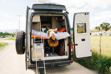 Positive schwarze Frau mit Afrofrisur liegt kopfüber auf dem Bett im Wohnwagen und surft auf dem Handy an einem sonnigen Sommertag - ADSF29790