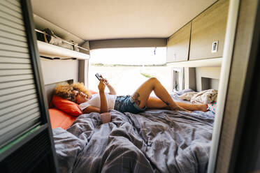 Anonyme Frau mit Afrofrisur liegt kopfüber auf dem Bett im Wohnwagen und surft auf dem Handy an einem sonnigen Sommertag - ADSF29792