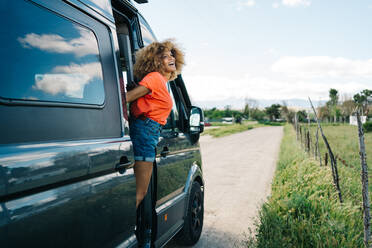 Fröhliche Afroamerikanerin steht in der Tür eines Wohnwagens und genießt die Landschaft - ADSF29798