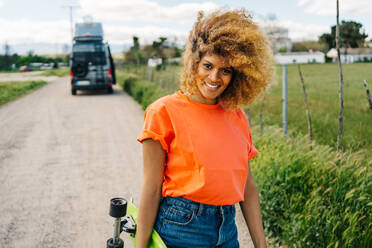 Glückliche afroamerikanische Frau, die ein Longboard trägt und lächelnd in die Kamera schaut, während sie auf einer Landstraße in der Nähe eines Vans im Sommer spazieren geht - ADSF29799