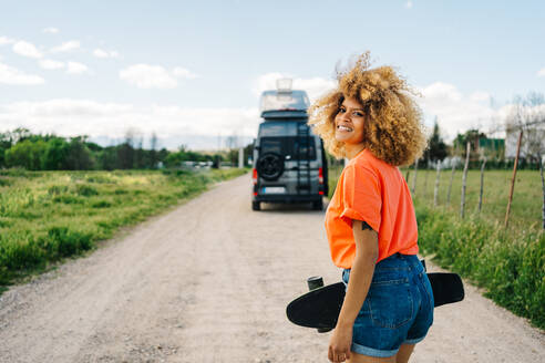 Happy African American female carrying longboard and looking away with smile while walking on countryside road near van in summer - ADSF29800
