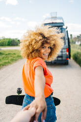 Glückliche afroamerikanische Frau mit Longboard, die lächelt und in die Kamera schaut, während sie einen Erntefreund während einer Autoreise zum Van führt - ADSF29801