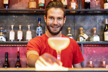 Fröhlicher Barmann, der ein Glas mit einem alkoholischen Cocktail auf dem Tresen einer Bar serviert und in die Kamera schaut - ADSF29804