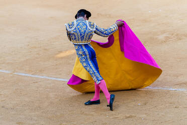 Rückenansicht eines anonymen Toreros in heller Tracht und mit Umhang, der während des Corrida-Festes auf einer sandigen Stierkampfarena auftritt - ADSF29809