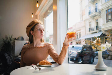 Französin mit Baskenmütze sitzt am Tisch in einem Café mit einem aromatischen Glas Kaffee und einem frisch gebackenen Croissant - ADSF29834