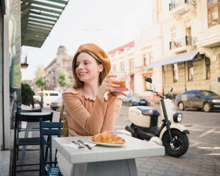 Französin mit Baskenmütze sitzt am Tisch in einem Café mit einem aromatischen Glas Kaffee und einem frisch gebackenen Croissant - ADSF29838