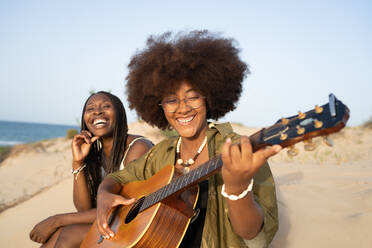 Fröhliche junge afroamerikanische Freundinnen spielen Gitarre, während sie am Strand sitzen und die Sommerferien genießen - ADSF29839